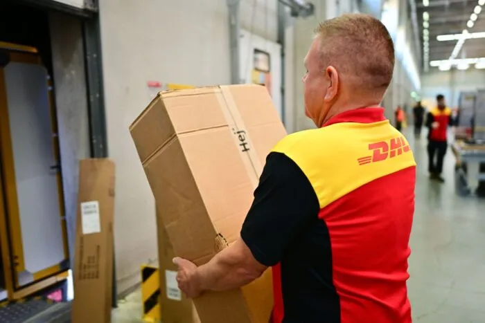 Ein Paketbote schleppt in einem DHL-Standort ein schweres Paket. (Archivbild)