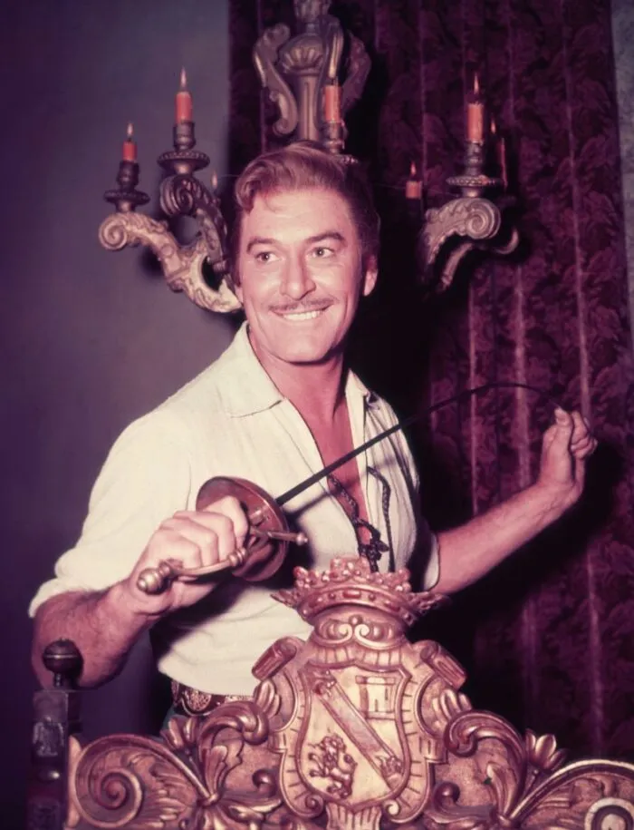 Um 1945: Porträt des in Australien geborenen Schauspielers Errol Flynn (1909–1959), der lächelnd hinter einem Stuhl steht und mit einem Schwert spielt, am Set eines seiner Abenteuerfilme, 1940er Jahre. Foto: Hulton Archive/Getty Images